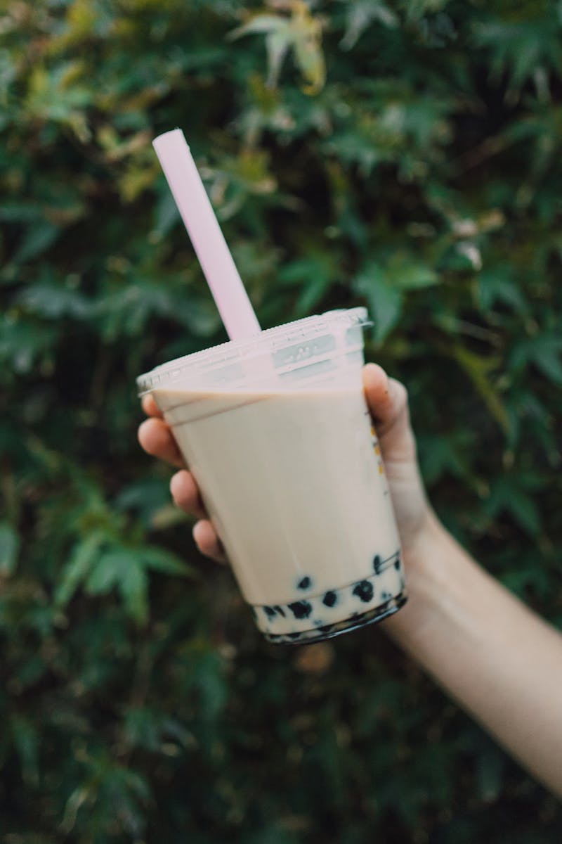 Close-up of a hand holding iced milk tea with boba pearls and straw against a leafy background.